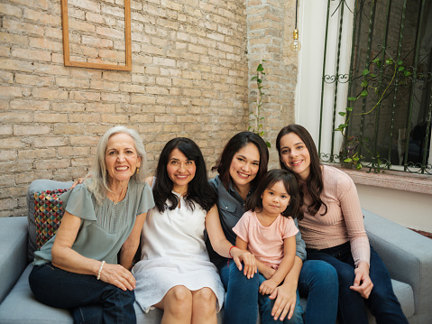 Beautiful intergenerational mexican women sitting together on a sofa, looking at the camera and smiling.
