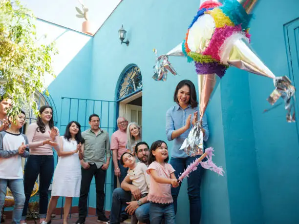 A happy mexican girl hitting a piñata with a stick next to her mother and family standing in the background looking at her.