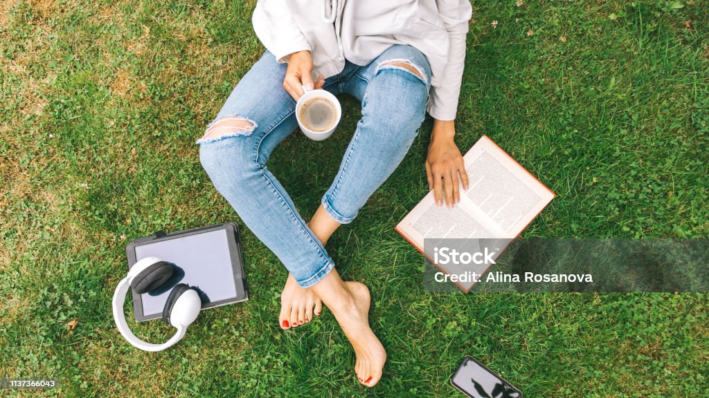 Young woman sitting on the grass drinking coffee and reading a book enjoys outdoor recreation. Woman drinking coffee and sitting with book on the grass Reading Stock Photo
