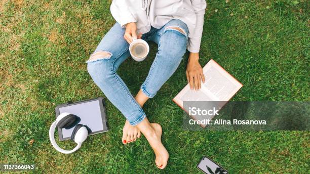 Young Woman Sitting On The Grass Drinking Coffee And Reading A Book Enjoys Outdoor Recreation Stock Photo - Download Image Now