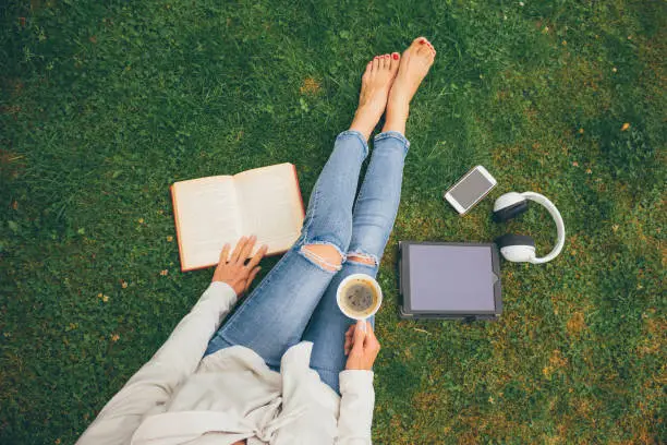 Photo of Top view of woman sitting in park (garden) on the green grass with smartphone, headphones, tablet, book and coffee in the hand