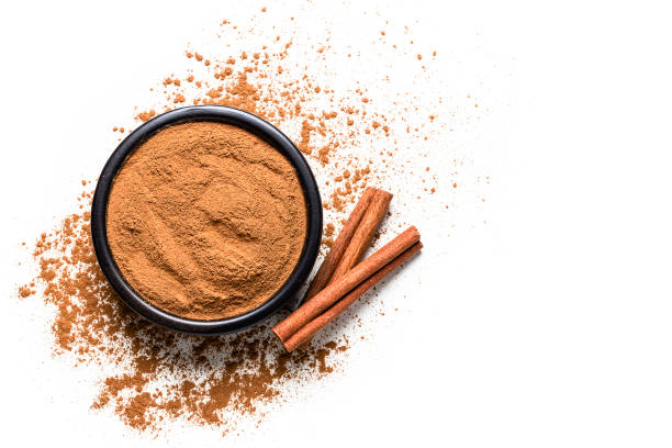 Spices: Top view of a black bowl filled with ground cinnamon isolated on white background. Cinnamon sticks are beside the bowl and ground cinnamon is scattered on the table. Useful copy space available for text and/or logo. Predominant colors are brown and white. High key DSRL studio photo taken with Canon EOS 5D Mk II and Canon EF 100mm f/2.8L Macro IS USM.