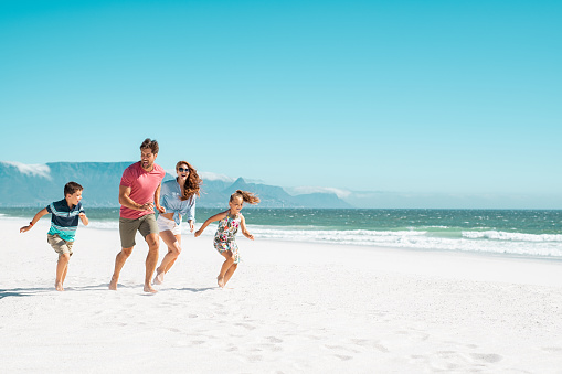 Happy young family running on the beach with copy space. Beautiful mother and laughing father enjoying summer vacation with son and daughter. Smiling parents with two children in casual having fun at seaside.