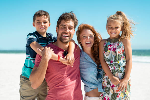 Happy family smiling at beach Portrait of happy family looking at camera at beach on a sunny day. Cheerful mother and father with cute daughter and son at sea during weekend. Smiling family with two children enjoying vacation at beach. beach holiday photos stock pictures, royalty-free photos & images