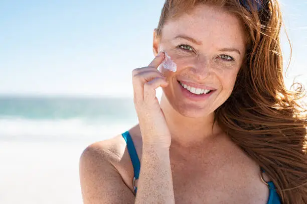 Smiling young woman applying sunscreen lotion on face at beach, with copy space. Beautiful mature woman with red hair enjoying summer at sea. Portrait of happy girl using sunblock on her delicate skin with freckles and looking at camera.