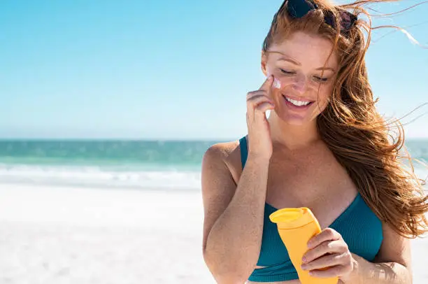 Beautiful young woman in blue bikini at beach applying sunscreen on face for protection on a sunny day. Mature woman with freckles and red hair enjoying summer holiday while applying suntan lotion. Portrait of smiling lady with healthy skin.