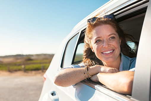 Smiling young woman peeking out of car window. Cheerful girl with freckles enjoying breeze from running car. Mature woman with red hair in the wind leans out of the car window and looking straight with copy space.