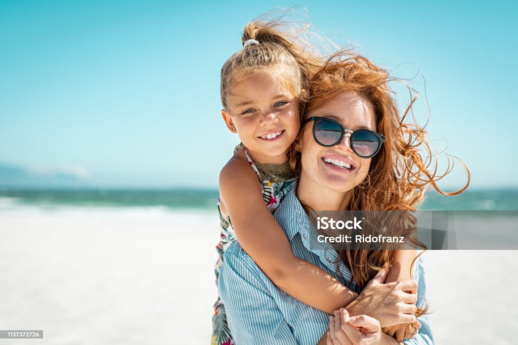 Mother with daughter at beach Smiling mother and beautiful daughter having fun on the beach. Portrait of happy woman giving a piggyback ride to cute little girl with copy space. Portrait of happy blonde kid embracing her mom wearing spectacles at beach during summer vacation. Beach Stock Photo