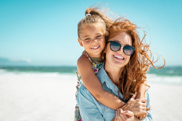 Mother with daughter at beach Smiling mother and beautiful daughter having fun on the beach. Portrait of happy woman giving a piggyback ride to cute little girl with copy space. Portrait of happy blonde kid embracing her mom wearing spectacles at beach during summer vacation. daughter photos stock pictures, royalty-free photos & images