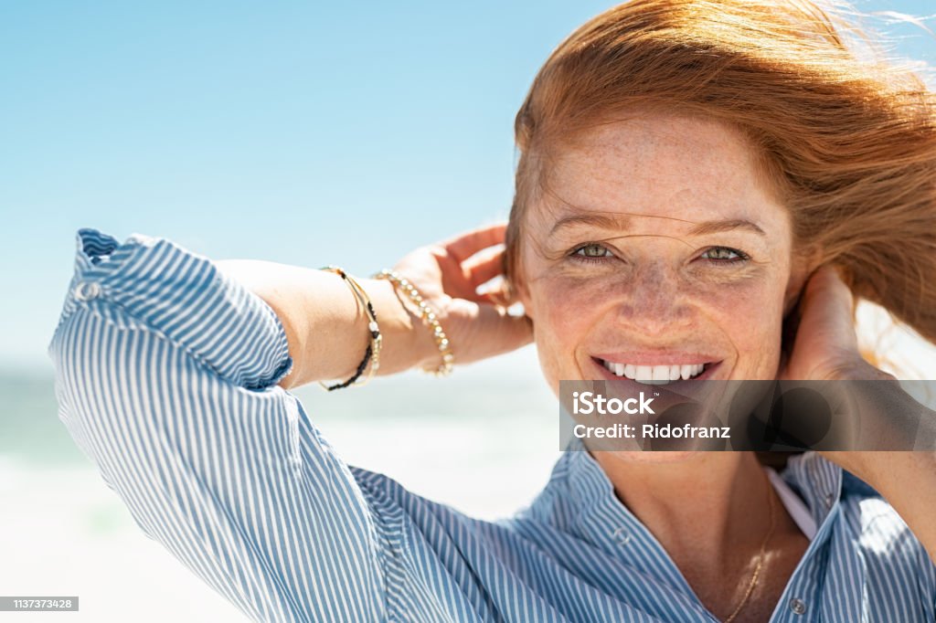 Smiling mature woman at beach Portrait of beautiful mature woman with wind fluttering hair. Closeup face of healthy young woman with freckles relaxing at beach. Cheerful lady with red hair and blue blouse standing at seaside enjoying breeze looking at camera. Women Stock Photo