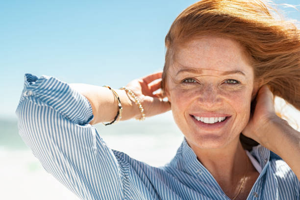 Smiling mature woman at beach Portrait of beautiful mature woman with wind fluttering hair. Closeup face of healthy young woman with freckles relaxing at beach. Cheerful lady with red hair and blue blouse standing at seaside enjoying breeze looking at camera. 40 44 years stock pictures, royalty-free photos & images