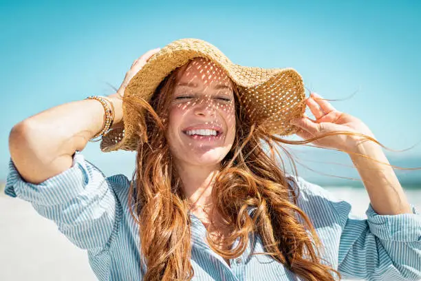 Closeup face of mature woman wearing straw hat enjoying the sun at beach. Happy young woman smiling during summer vacation at sea. Portrait of beautiful lady relaxing at beach while holding large brim for the wind.