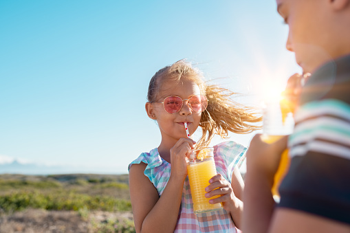 Portrait of little girl drinking orange juice in a glass with straw. Rear view of brother drinking with sister at outdoor park. Boy and cute girl with sunglasses suck from the straw a fresh pineapple juice outdoor with copy space.