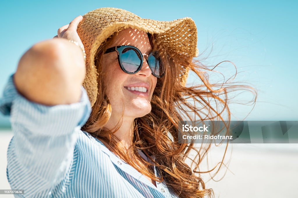 Mature woman with beach hat and sunglasses Side view of beautiful mature woman wearing sunglasses enjoying at beach. Young smiling woman on vacation looking away while enjoying sea breeze wearing straw hat. Closeup portrait of attractive girl relaxing at sea. Women Stock Photo