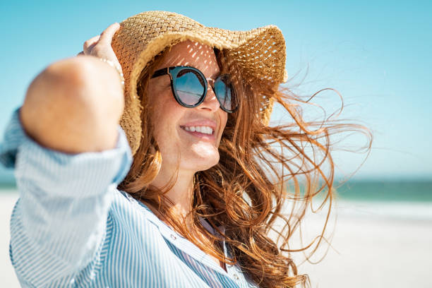 Mature woman with beach hat and sunglasses Side view of beautiful mature woman wearing sunglasses enjoying at beach. Young smiling woman on vacation looking away while enjoying sea breeze wearing straw hat. Closeup portrait of attractive girl relaxing at sea. sunbathing stock pictures, royalty-free photos & images