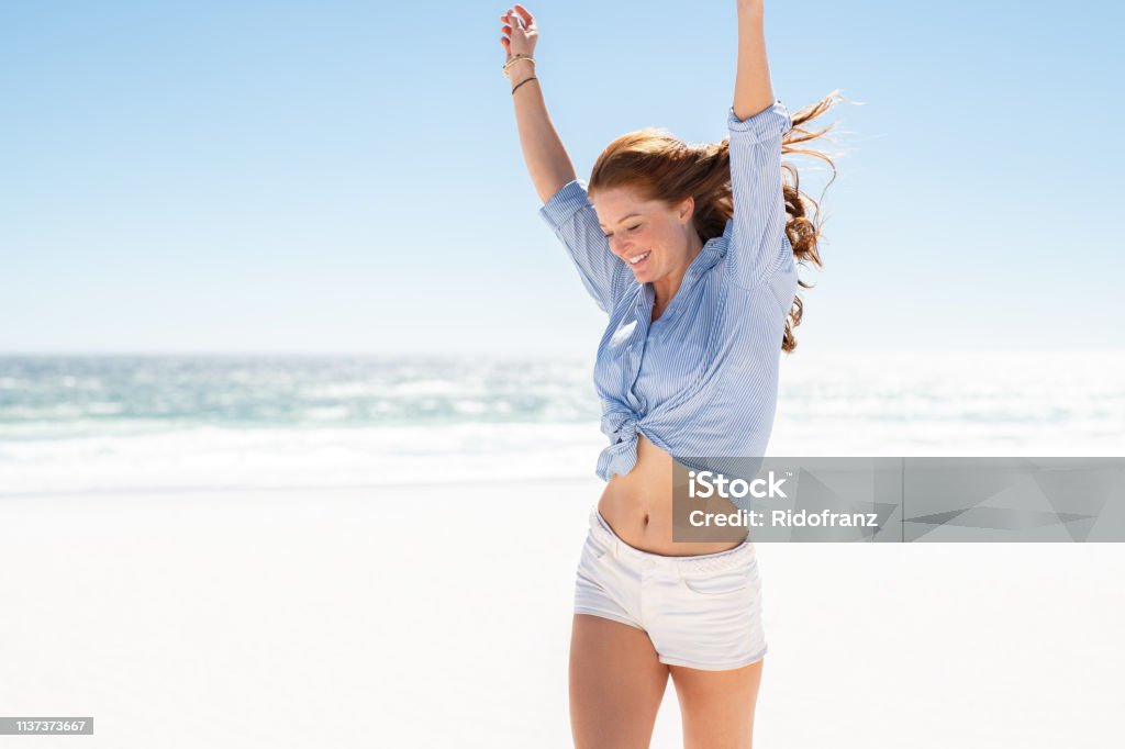 Smiling woman enjoying the beach Happy mature woman in blue blouse and white shorts enjoying tropical beach vacation. Smiling young woman having fun on her vacation at sea. Joyful lady with red hair enjoying freedom with outstreched arms, jumping on beach with copy space. Women Stock Photo