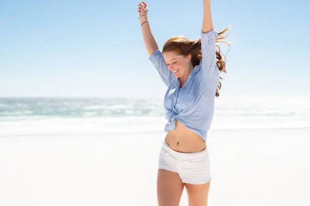 Happy mature woman in blue blouse and white shorts enjoying tropical beach vacation. Smiling young woman having fun on her vacation at sea. Joyful lady with red hair enjoying freedom with outstreched arms, jumping on beach with copy space.