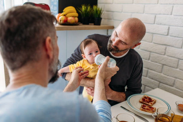 LGBTQ Family - Gay Dads Gay dads with their adopted baby by the table. Having breakfast and giving water to baby. gay daddy stock pictures, royalty-free photos & images