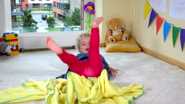 Mischievous toddler girl hide under yellow plaid blanket on floor