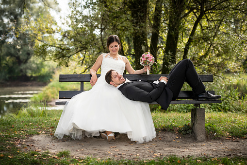 Just married bride sitting on a bench at an idyllic place at a lake. Her groom is lying on her looking happy at the camera.