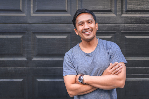 Portrait of a middle aged man standing in front of garage doors. He is happy and he is smiling. He is wearing casuals in yard.
