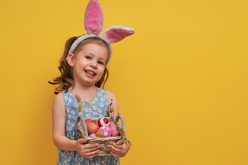 Cute little child wearing bunny ears on Easter day. Girl with painted eggs on bright yellow background.