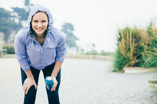 Cheerful jogger catching breath. Young woman in sports hoodie and tights leaning forward, holding water bottle and smiling. Jogging or stress relief concept