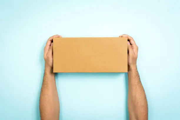 Photo of Concept of delivery and storage. Man showing a cardboard box with empty space. Only hands holding a closed box on blue background.
