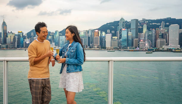 pares asiáticos felizes que apreciam um vidro de champagne com a opinião da skyline de hong kong - wine port - fotografias e filmes do acervo