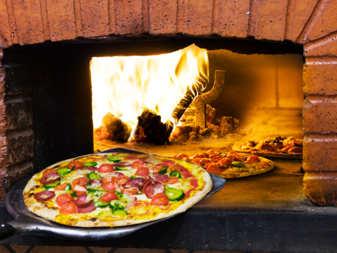 An Italian pizza is being removed from a wood burning oven on a large pizza spatula.  There are two more pizza's cooking in the oven.  The ingredients on the pizza's are a combination of different meats, vegetables and cheeses.  Fire is visible towards the back left side of the oven.