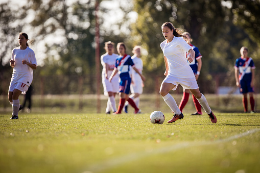 Teenage soccer player passing her opponents while running with a ball on playing filed.