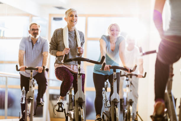 Group of active seniors having exercise class with an instructor in a gym. Happy mature athletes exercise on exercises bikes while having sports training with their coach. Focus is on woman with short hair. spin bike stock pictures, royalty-free photos & images