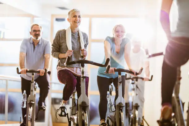 Happy mature athletes exercise on exercises bikes while having sports training with their coach. Focus is on woman with short hair.