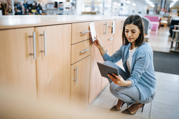 Young woman checking price tag while using digital tablet at furniture store. Female worker using touchpad and checking price tags while working at furniture store. compare price online stock pictures, royalty-free photos & images