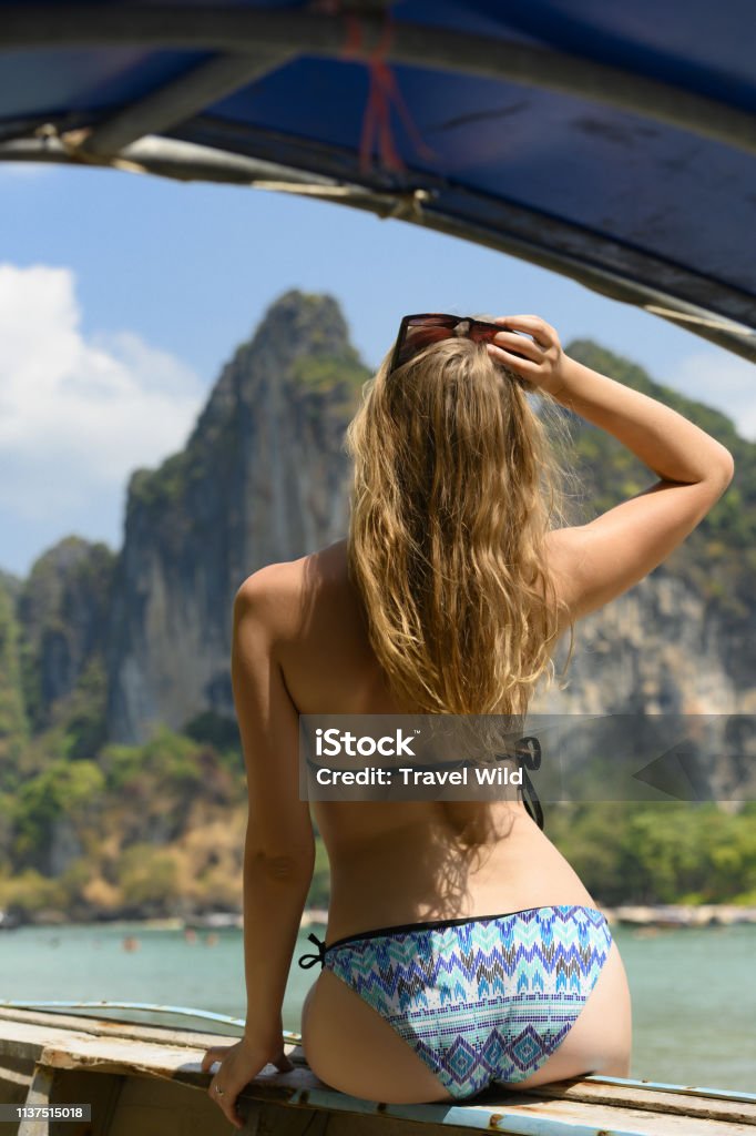 An unidentified young blonde girl, wearing bikini, is admiring the landscape sitting on a wooden boat in Raiwai Beach. Photo framed by a long tail boat. Ao Nang, Krabi, Thailand. Admiration Stock Photo