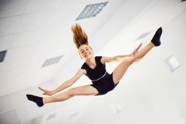 Split jump. Young female dancer jumped and made a split in the air at dance class. She's smiling and looking at camera. legs apart stock pictures, royalty-free photos & images