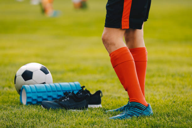 jambes d'un garçon de joueur de football dans des cales de bottes avec une bille et un rouleau de mousse sur la pelouse verte du stade. équipement de football gros plan - foot massage photos et images de collection