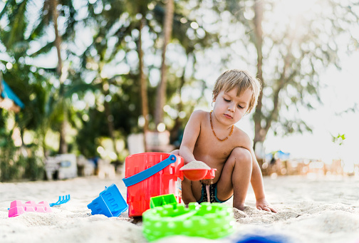 Small boy sitting in sand at the beach and playing with his toys.