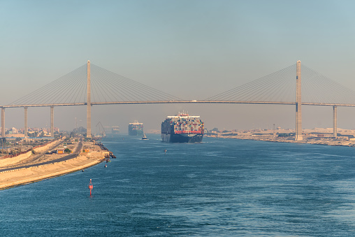 El Qantara, Egypt - November 5, 2017: Container vessel ships passing Suez Canal in the sandy haze in Egypt. The Mubarak Peace Bridge is a road bridge crossing the Suez Canal at El-Qantara.