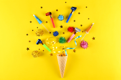 Birthday celebration attributes. Party horns, colorful confetti, candles ice cream cone, cocktail umbrellas & other items on bright yellow background. Close up, top view, copy space, flat lay.