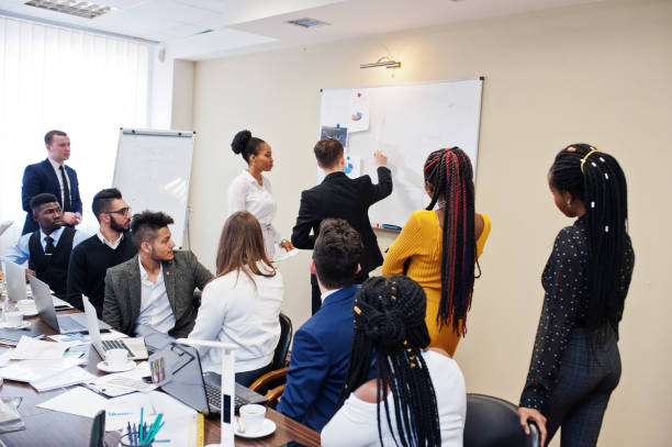 istock Mixed race business coach presenting report standing near whiteboard pointing on sales statistic shown on diagram and chart teach diverse company members gathered together in conference room. 1137533414