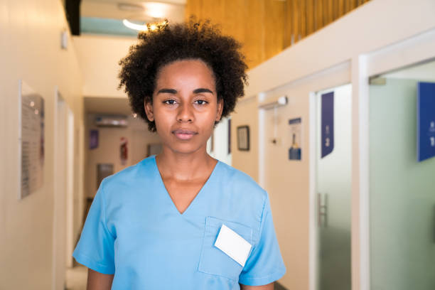 Portrait of young healthcare worker standing with blank expression. Confident female nurse is in blue scrubs. She is in corridor at hospital.