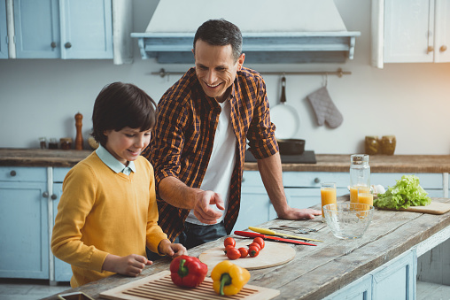 Man and boy preparing food in the kitchen. Adult pointing at vegetable on the table. They are smiling