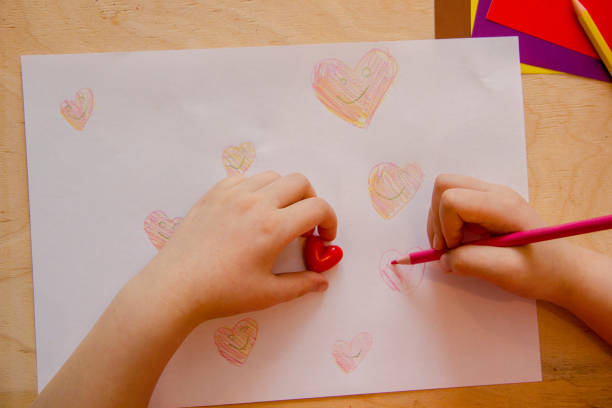 istock Child drawing pencil on white paper sheet with wooden texture background, top view. Artwork workplace with creative accessories.Close-up of child hands drawing a red heart 1137568229