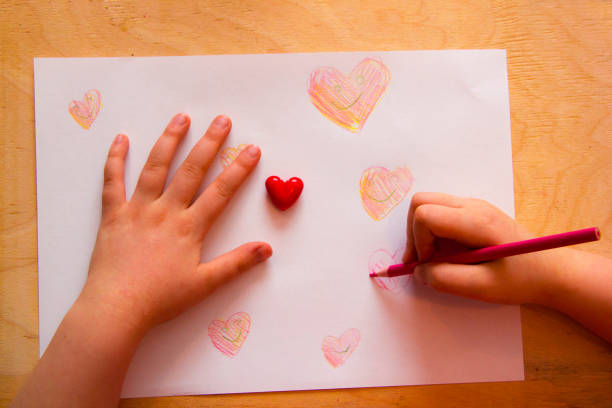 istock Child drawing pencil on white paper sheet with wooden texture background, top view. Artwork workplace with creative accessories.Close-up of child hands drawing a red heart 1137568307