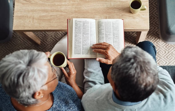 Let's turn the page High angle shot of a relaxed elderly couple reading from the bible while drinking coffee at home during the day bible stock pictures, royalty-free photos & images