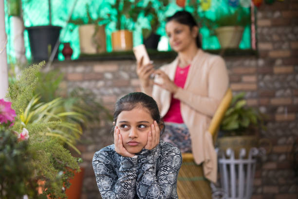 Bored girl with mother using mobile phone Bored daughter with mother using mobile phone in the background ignoring stock pictures, royalty-free photos & images