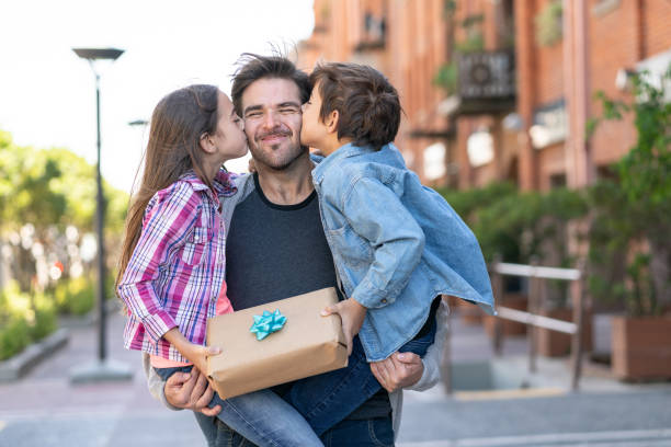Loving son and daughter kissing daddy on the cheek while he carries them both with a present for father's day
