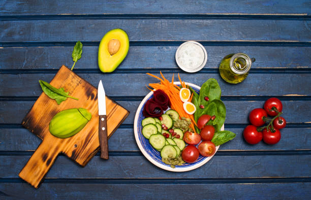istock Cooking. Preparing vegan salad with avocado beets carrots spinach tomatoes cucumber and sunflower seeds. Buddha detox bowl Recipe. Woman hands with knife and fork. Blue background. Top view, flat copy space. 1137616435