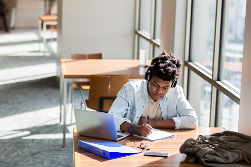 African American teenage boy writes something in a notebook while studying in the campus library. An open laptop is on the table. He is wearing wireless headphones.